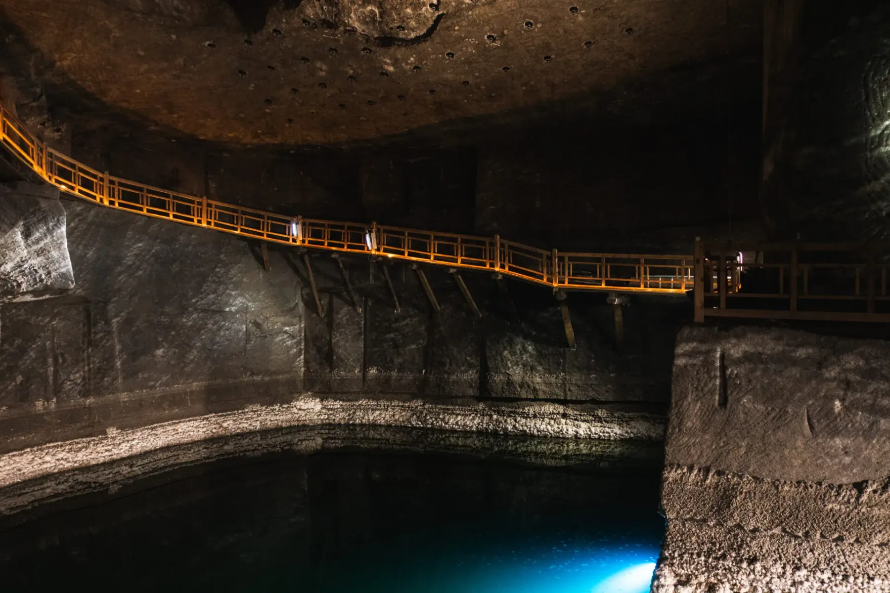 Ein beleuchteter Holzsteg verläuft entlang der Höhlenwand über einem unterirdischen Salzsee im Salzbergwerk Wieliczka.