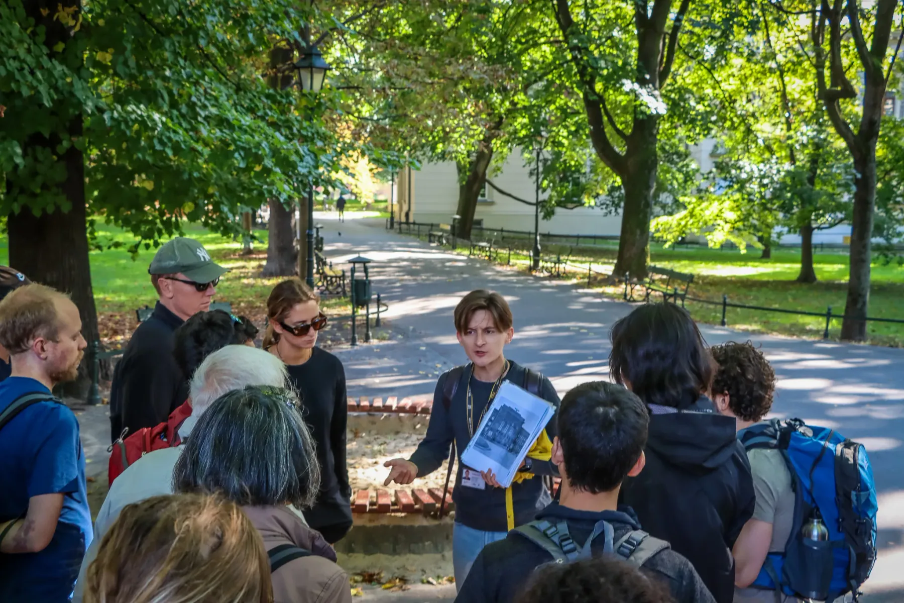 Eine Reiseführerin zeigt einer Gruppe von Personen auf einem Weg in einem sonnigen Park in Krakau historische Fotos.