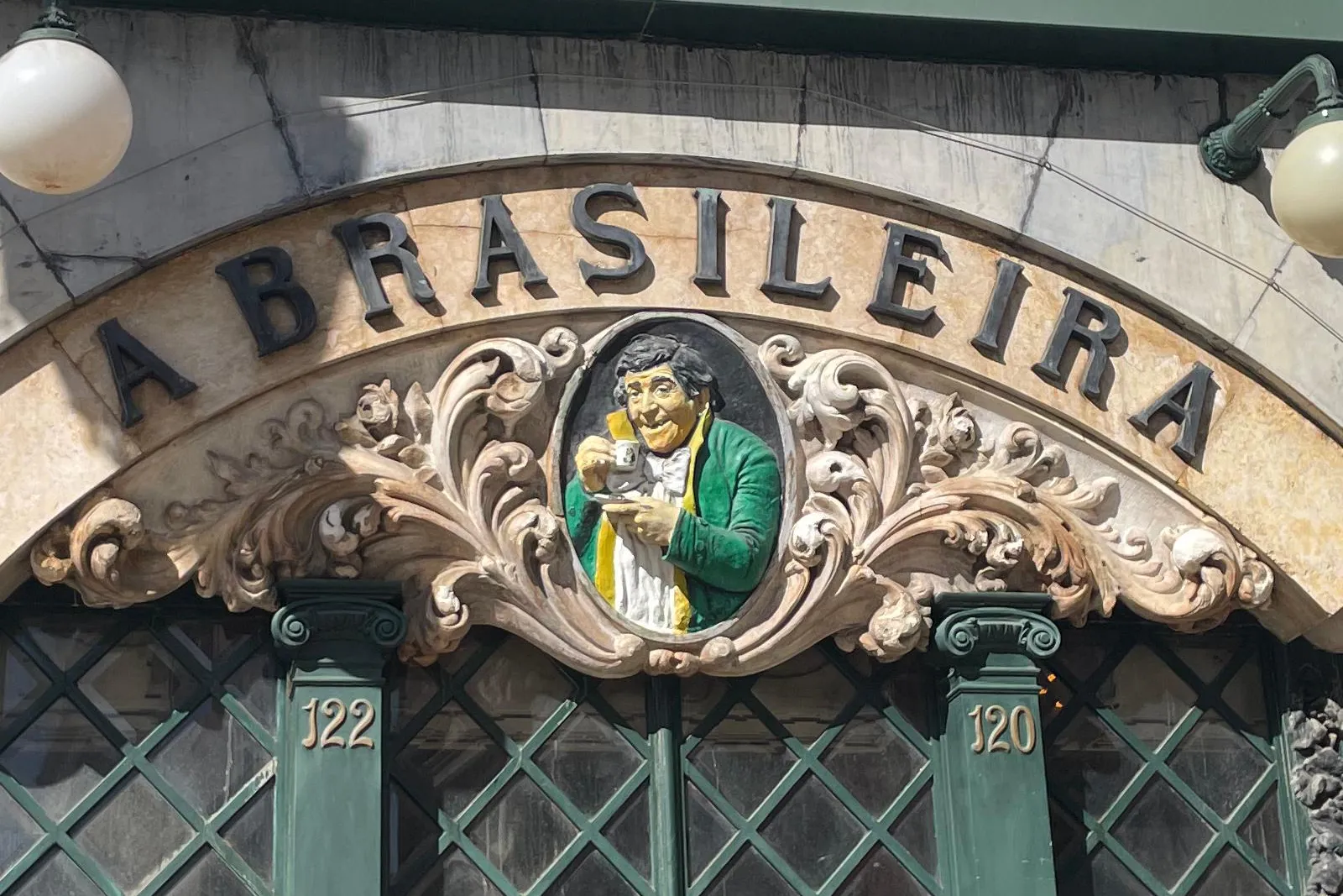 Fassade des Café A Brasileira in Lissabon, mit bogenförmigem Schild, zentralem Relief eines Mannes mit Tasse und grün gera...