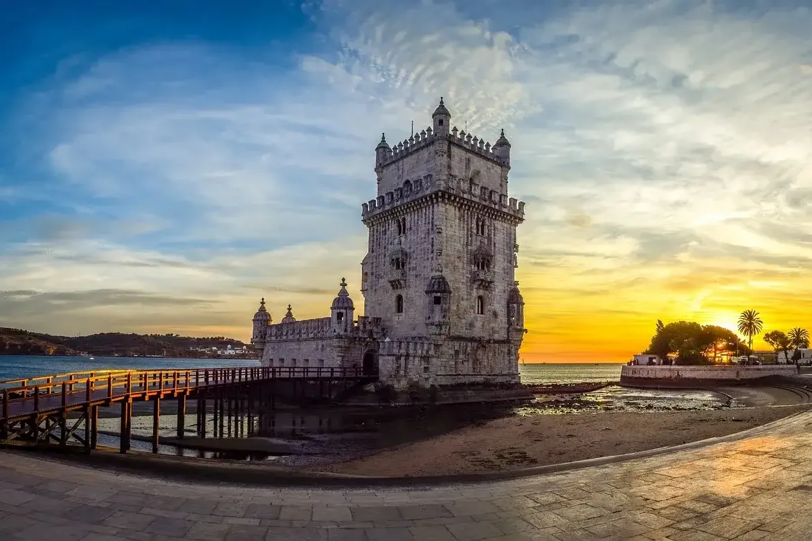 Historischer Turm von Belém in Lissabon bei Sonnenuntergang, mit Holzsteg über dem Tejo und orange-blauem Himmel.