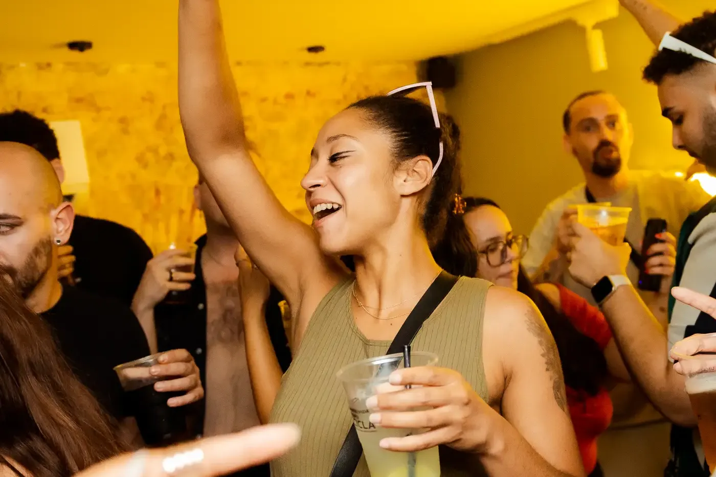 Gruppe tanzender Menschen, die Getränke in einer warm beleuchteten Bar halten. Eine Frau im Vordergrund lächelt mit erhobe...