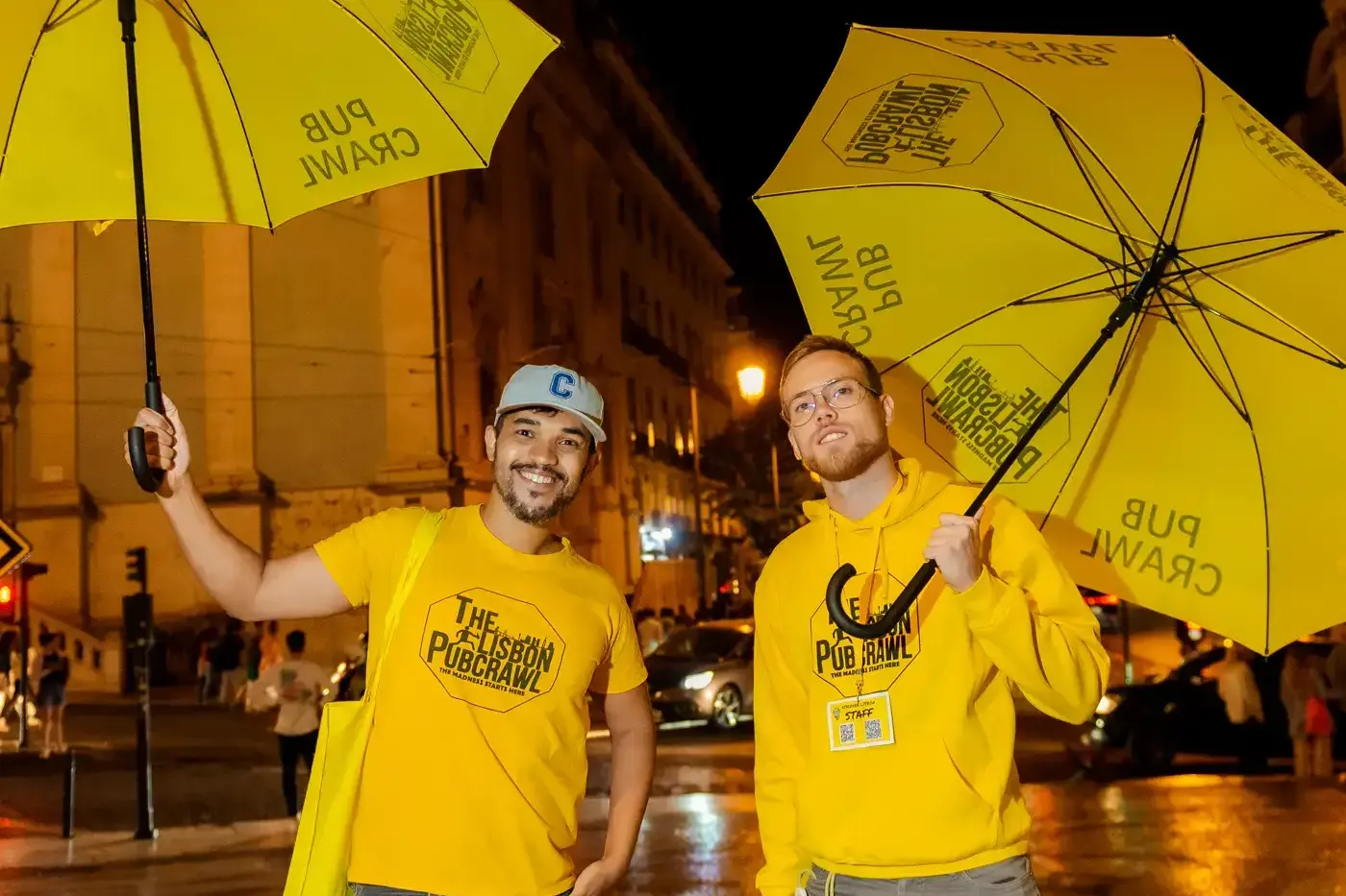 Zwei Männer in gelber 'The Lisbon Pubcrawl'-Kleidung mit gelben Regenschirmen stehen nachts auf nasser Straße in Lissabon.