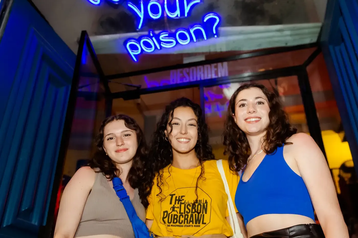 Drei junge Frauen lächeln vor einem blauen Neonschild mit der Aufschrift 'What's your poison?' über einem blauen Türrahmen.