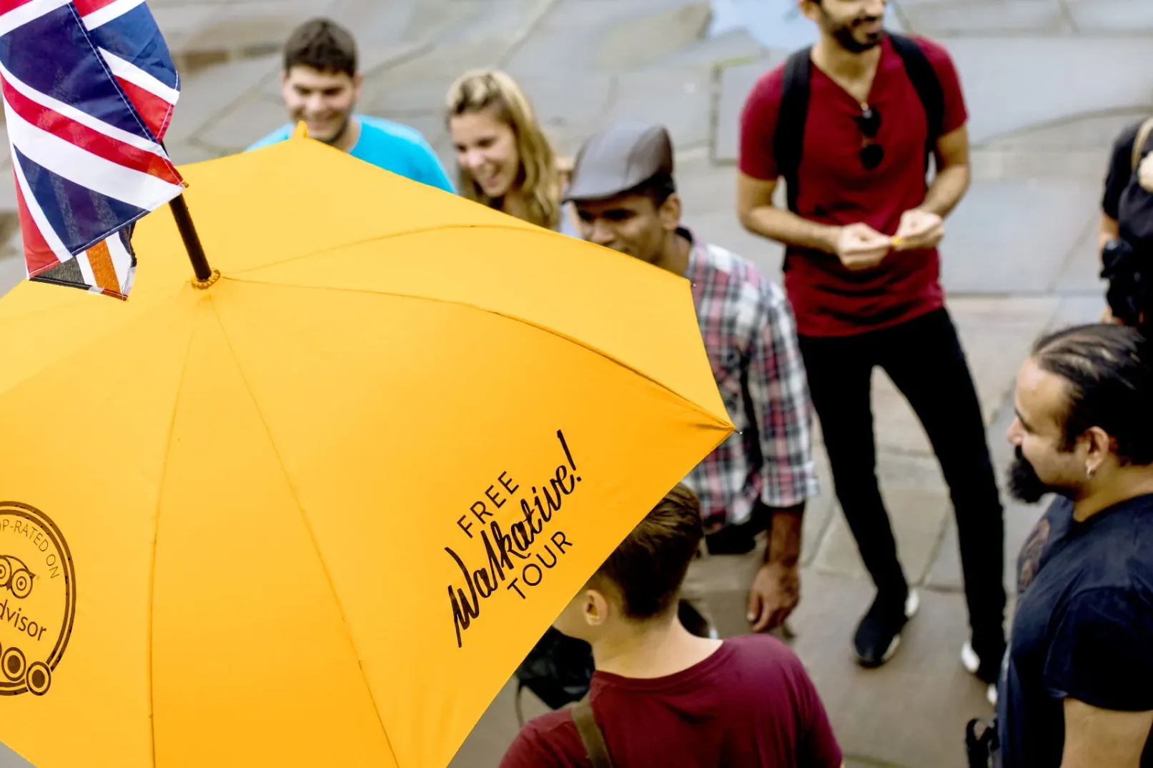 Eine vielfältige Gruppe von Menschen hört einem Reiseleiter zu, der einen gelben Walkative-Regenschirm mit kleiner Union J...