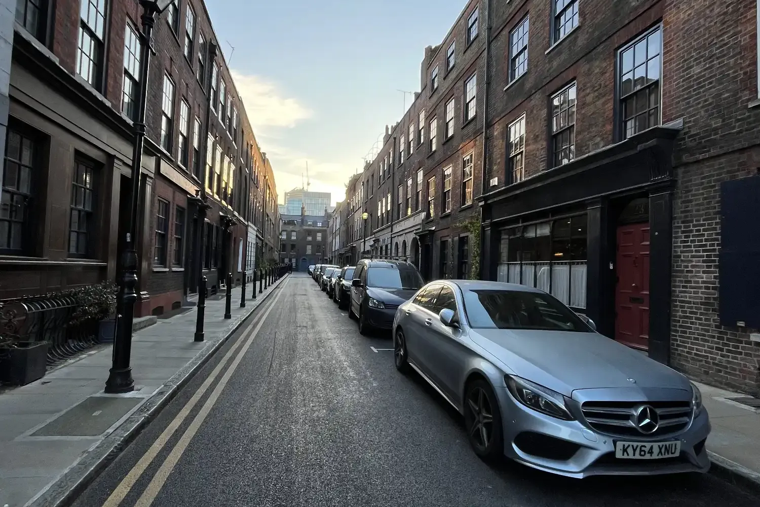 A narrow London street with cars parked between historic brick buildings at sunset.