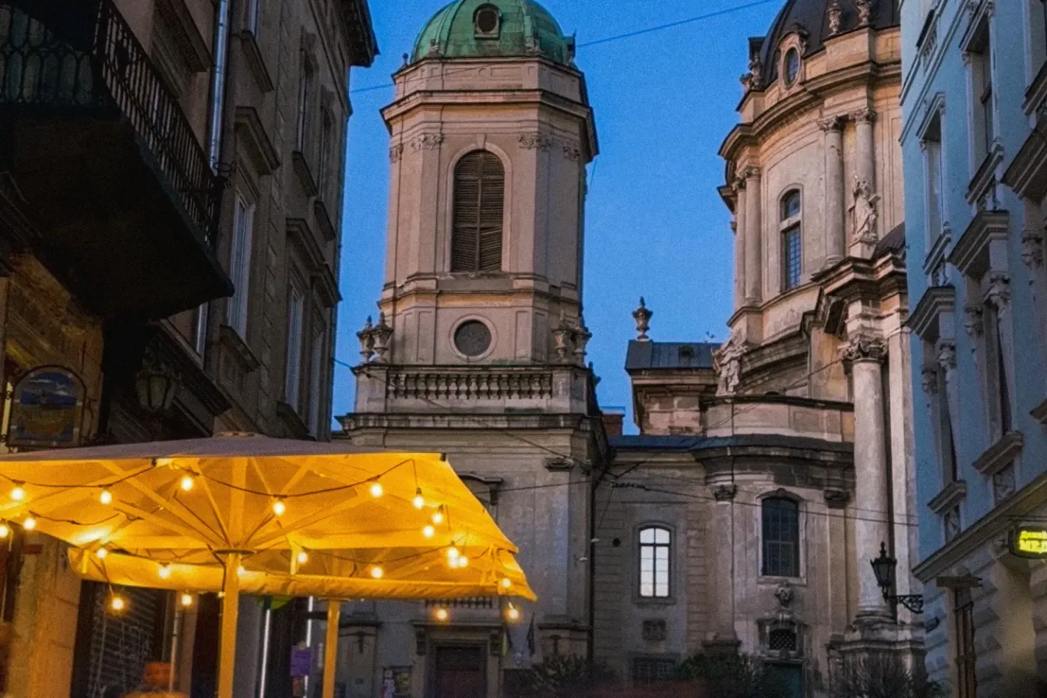 Ein belebtes Straßencafé in Lemberg bei Dämmerung, mit der Dominikanerkirche im Hintergrund unter einem tiefblauen Himmel.