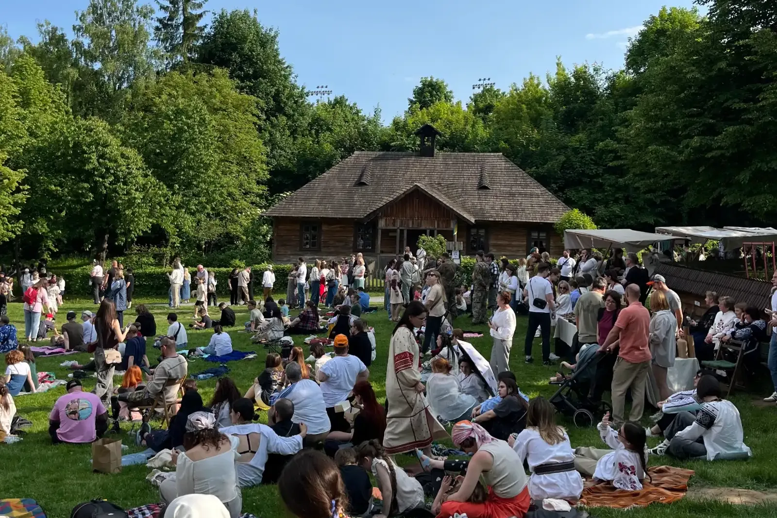 Eine große Menschenmenge versammelt sich an einem sonnigen Tag auf einer Wiese vor einem traditionellen Holzhaus.