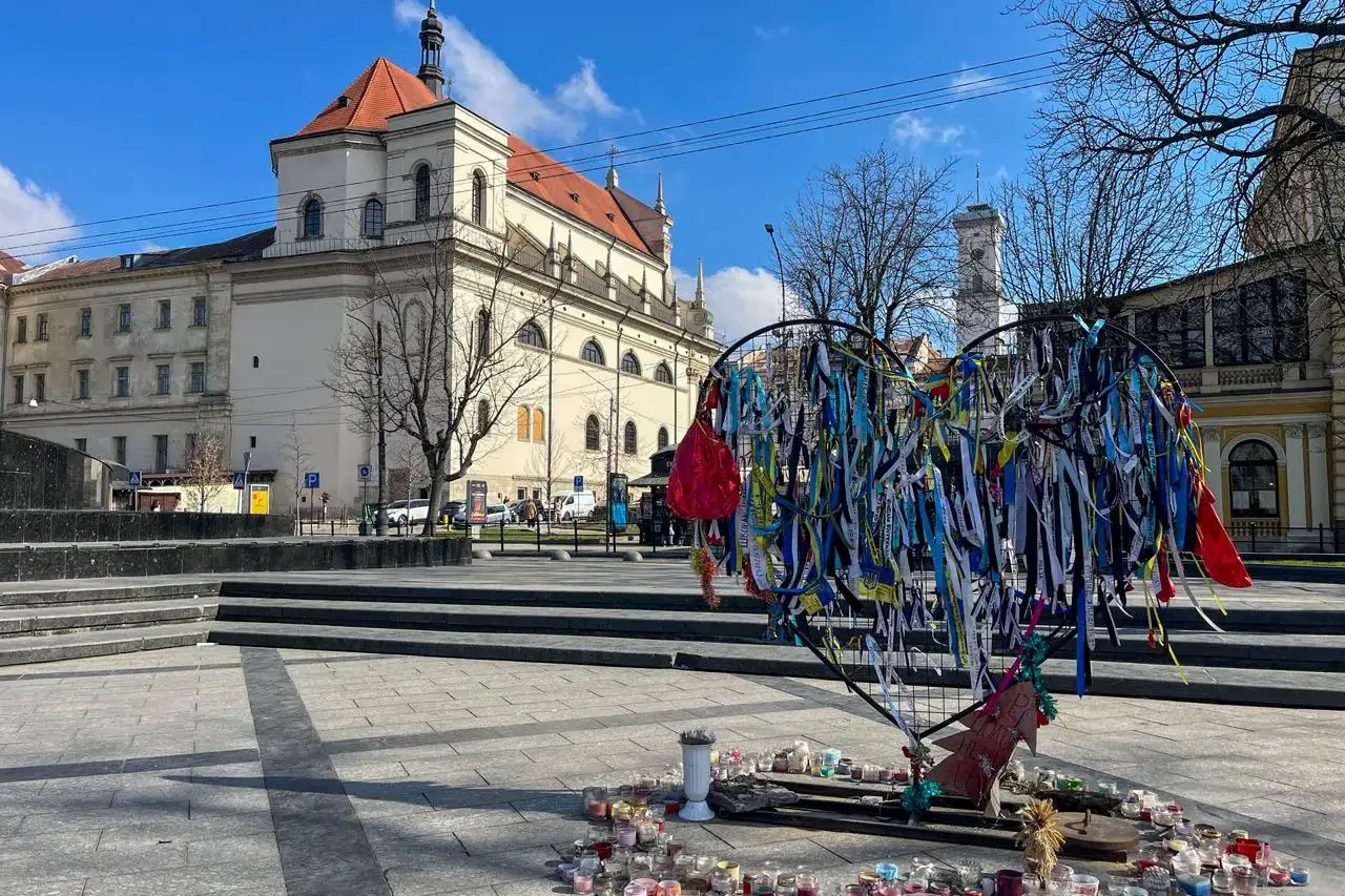 Herzförmiges Mahnmal mit blau-gelben Bändern und vielen Kerzen auf einem Platz in Lwiw, historisches Gebäude im Hintergrund.