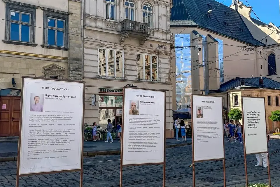 Four memorial stands with portraits and text on a cobblestone street in Lviv, with historic buildings and a church in the ...
