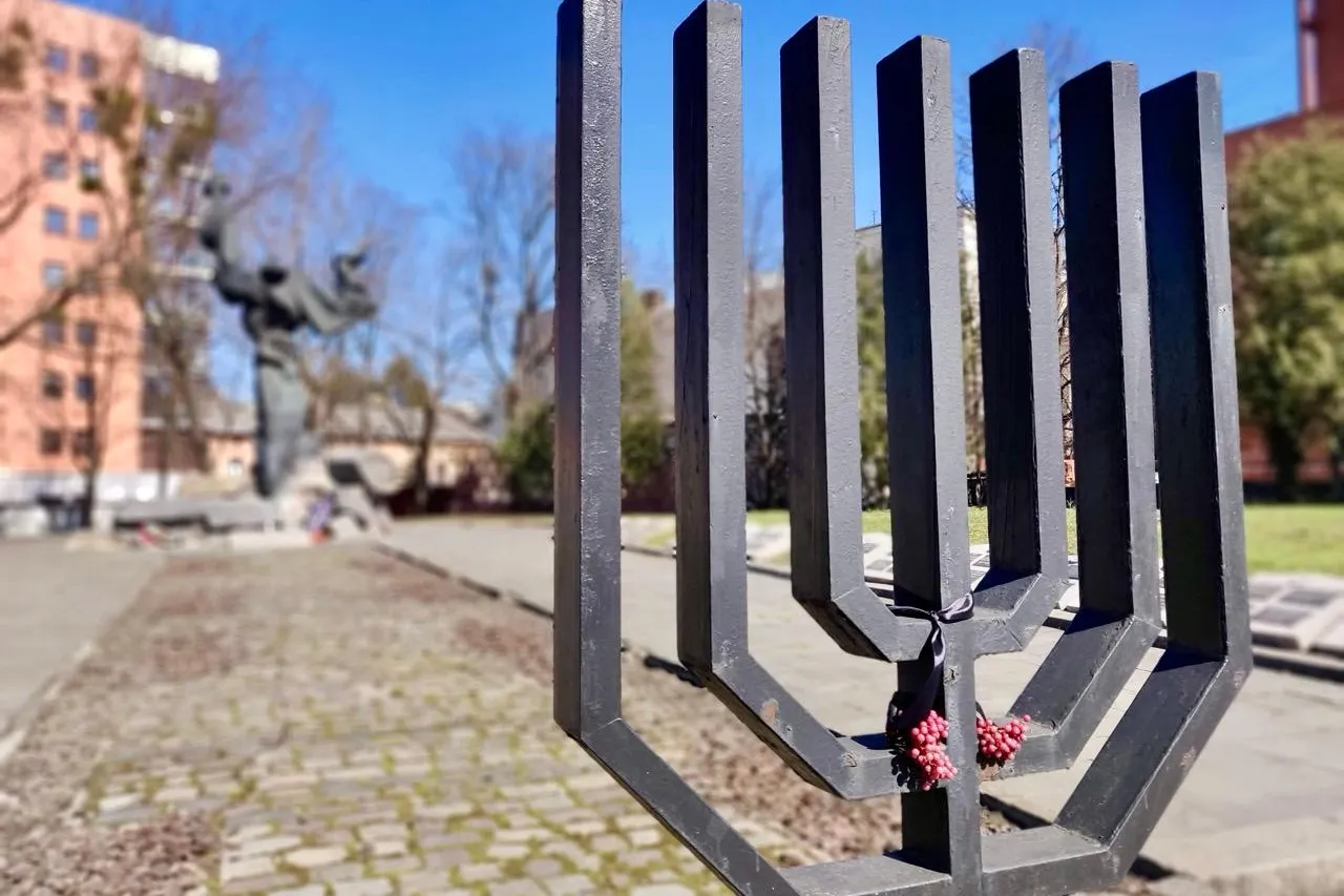 Black metal menorah sculpture with red berries tied to it, in front of a blurred abstract monument, building, and trees.