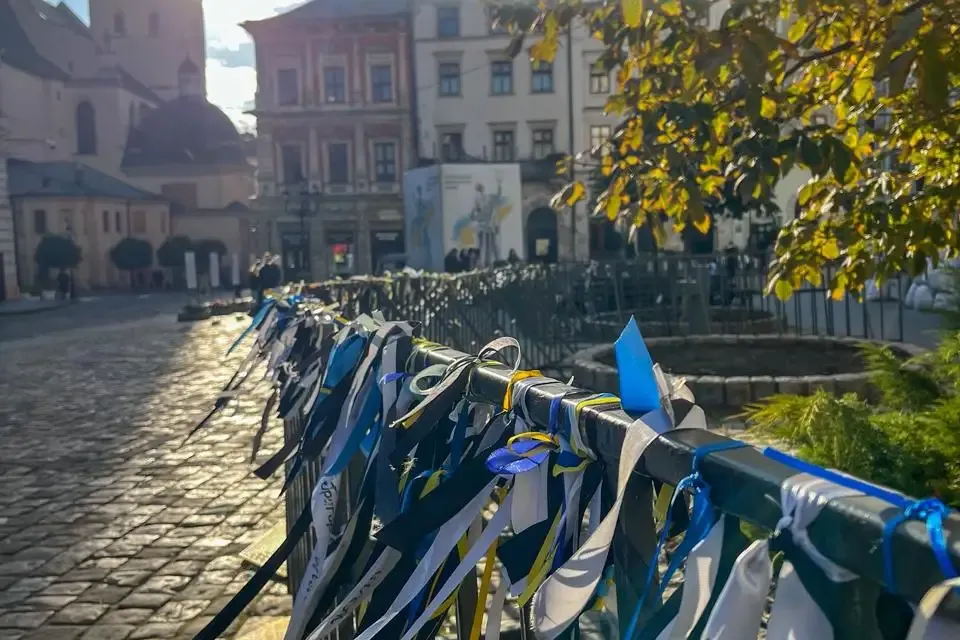 A fence along a cobblestone street in Lviv, adorned with many blue and yellow ribbons. St. Andrew's Church and historic bu...