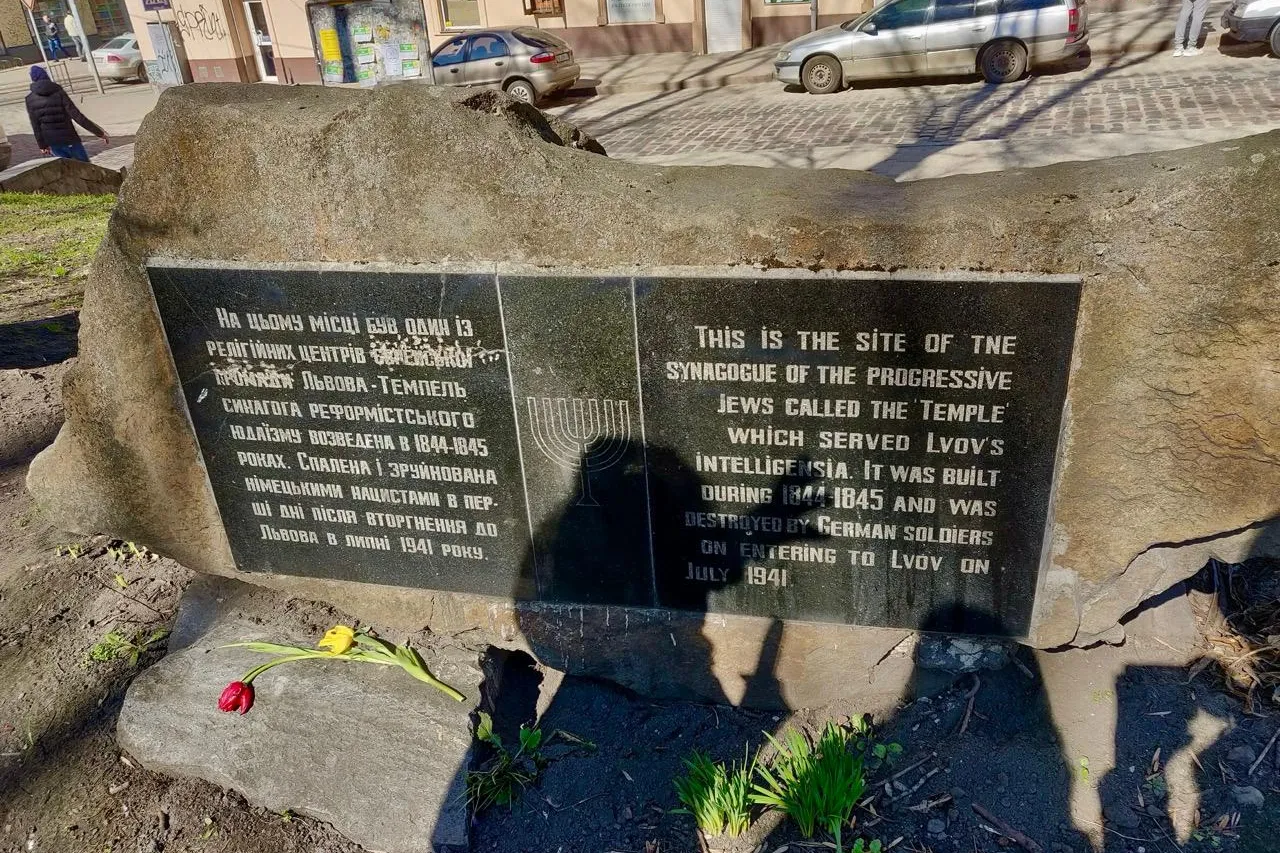 Stone monument with plaques in English and Ukrainian, marking the site of a destroyed synagogue. Two flowers lie at its ba...