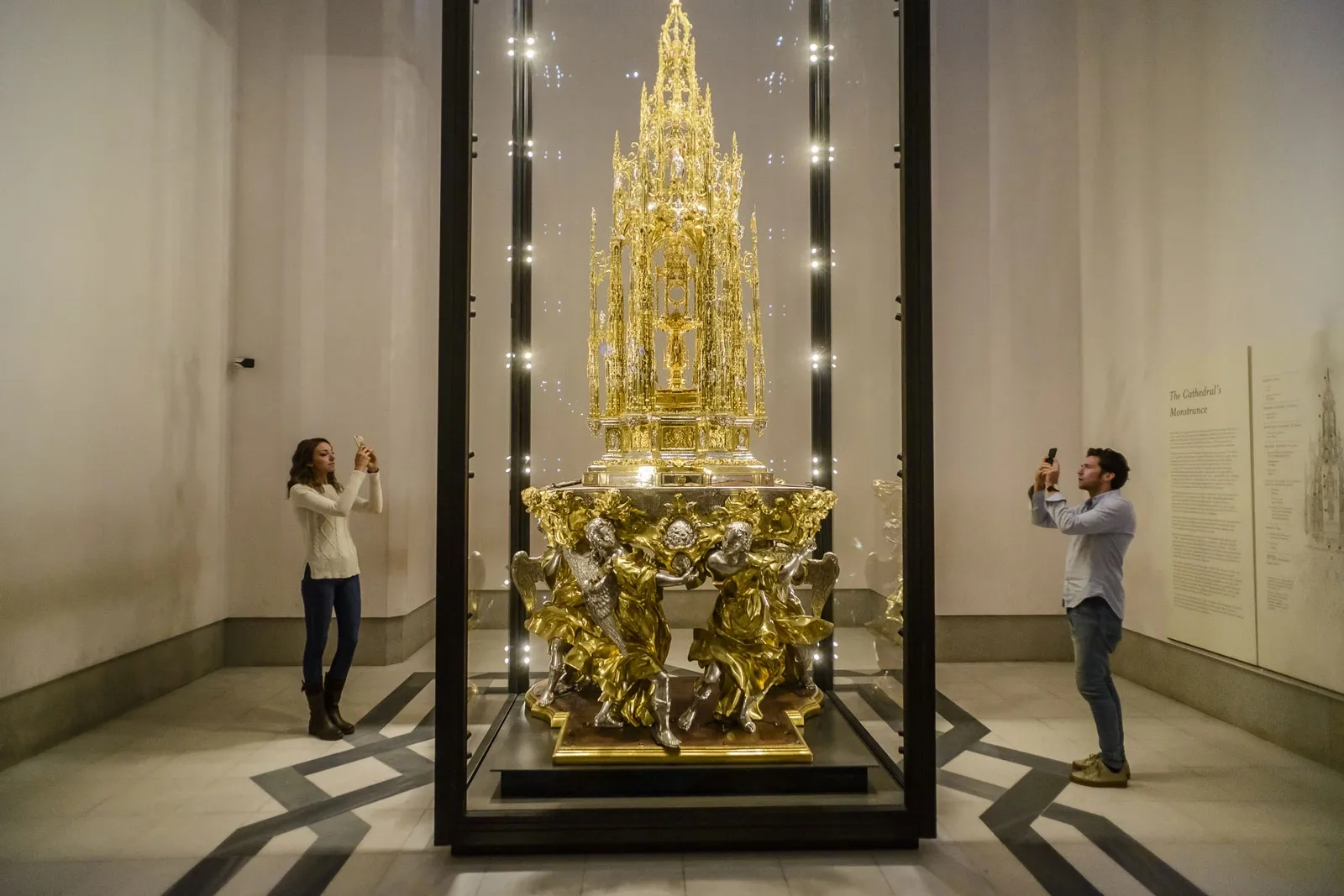 Eine reich verzierte, vergoldete Monstranz in einer Glasvitrine, die von zwei Besuchern mit ihren Handys fotografiert wird.