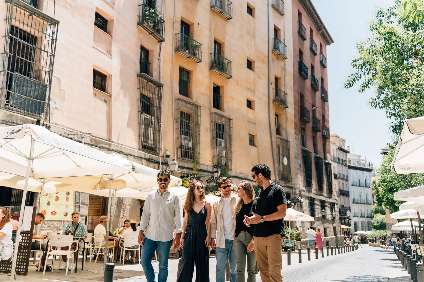 Eine Gruppe von fünf jungen Leuten spaziert durch eine sonnige Straße in Madrid mit Cafétischen und alten Gebäuden.
