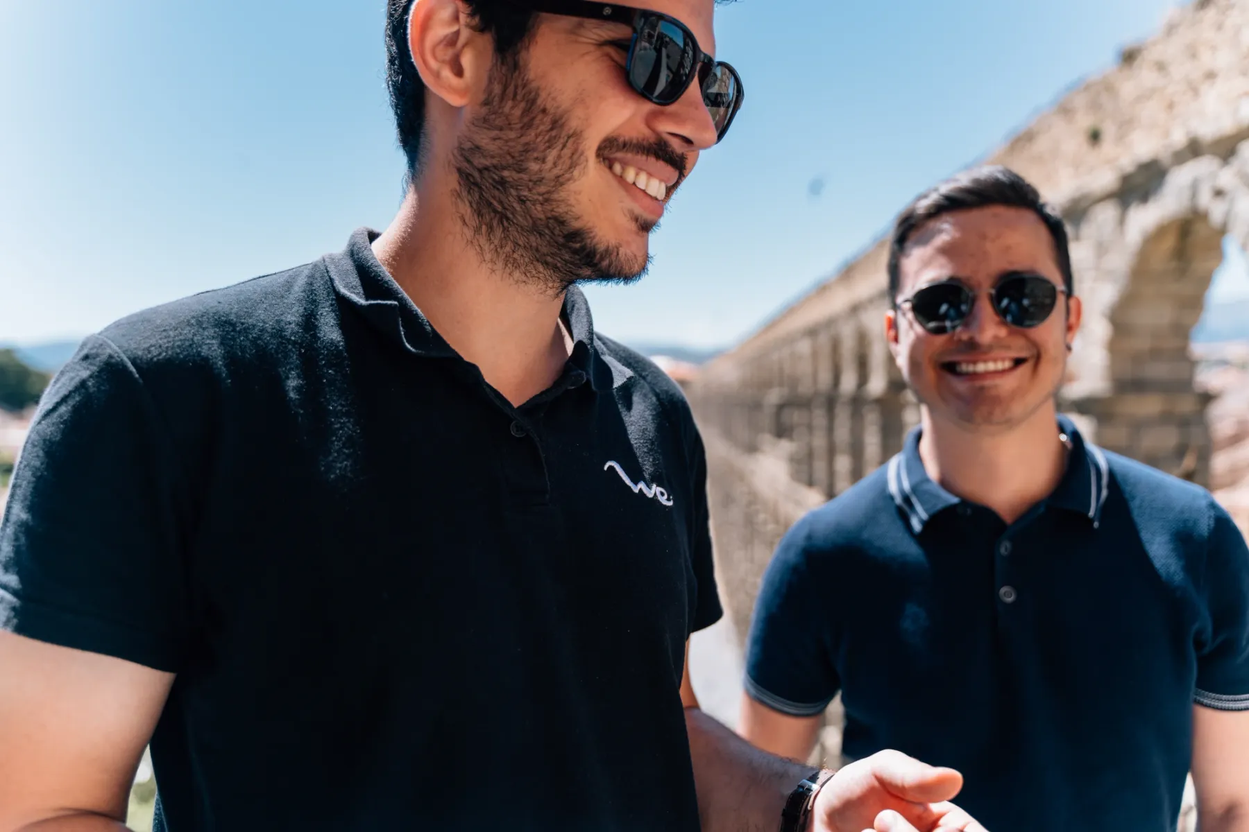 Zwei junge Männer mit Sonnenbrille stehen lächelnd vor dem Aquädukt von Segovia an einem sonnigen Tag.