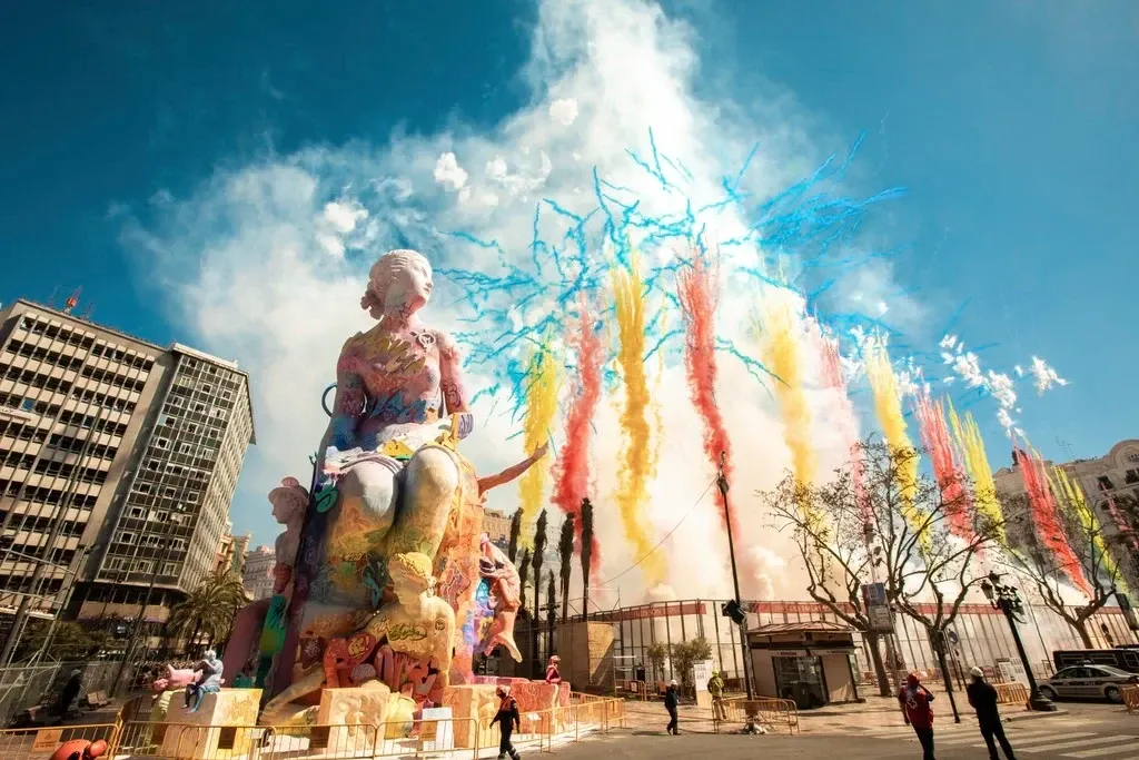 Una gran y colorida estatua de una 'falla' con un espectáculo de fuegos artificiales diurnos de humo de colores detrás de ...