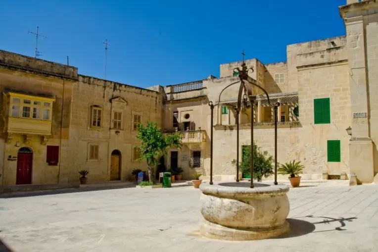 Plaza de piedra vacía con pozo histórico en primer plano. Edificios tradicionales con balcones y coloridas persianas rodea...