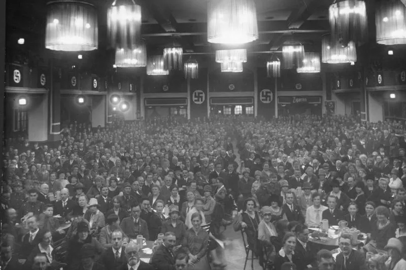 Foto en blanco y negro de un gran salón abarrotado de gente sentada en mesas. Banderas con esvásticas y letrero de 'Zigare...