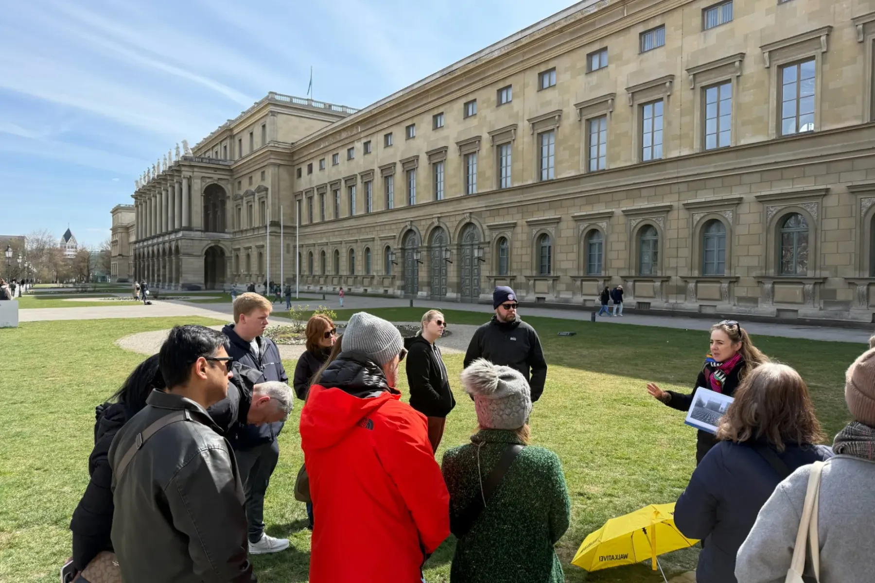 A tour guide with a yellow Walkative umbrella speaks to a group on a lawn in front of a large neoclassical building in Mun...