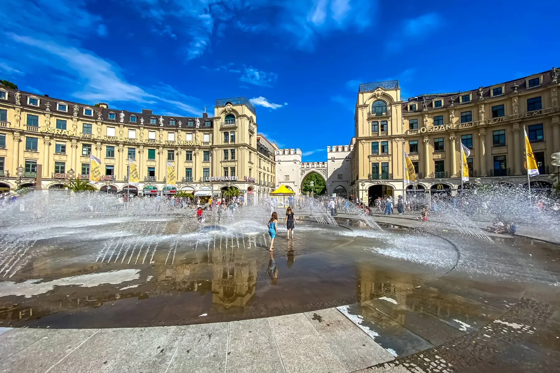 People enjoying the large fountain at Karlsplatz in Munich, with Karlstor archway and historic buildings in the background...