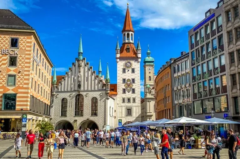 Zatłoczony Marienplatz w Monachium ze Starym Ratuszem i wieżą Nowego Ratusza pod błękitnym niebem.