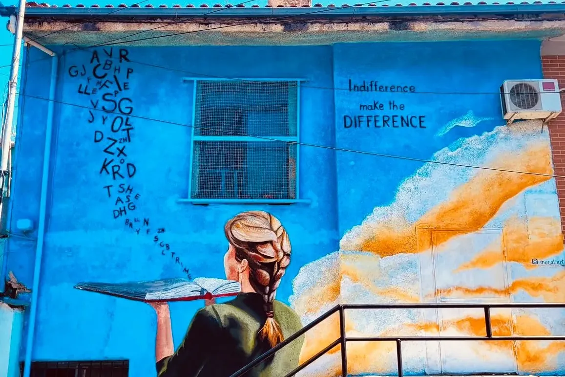 Mural na niebieskim budynku w Tiranie przedstawiający kobietę trzymającą otwartą książkę, z której wylatują litery, oraz n...