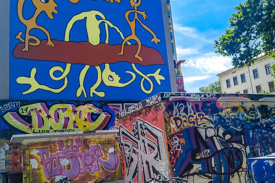 Blaue Gebäudewand mit großem gelb-roten abstrakten Wandgemälde. Darunter Wände mit bunten Graffiti, geparkte Fahrräder und...