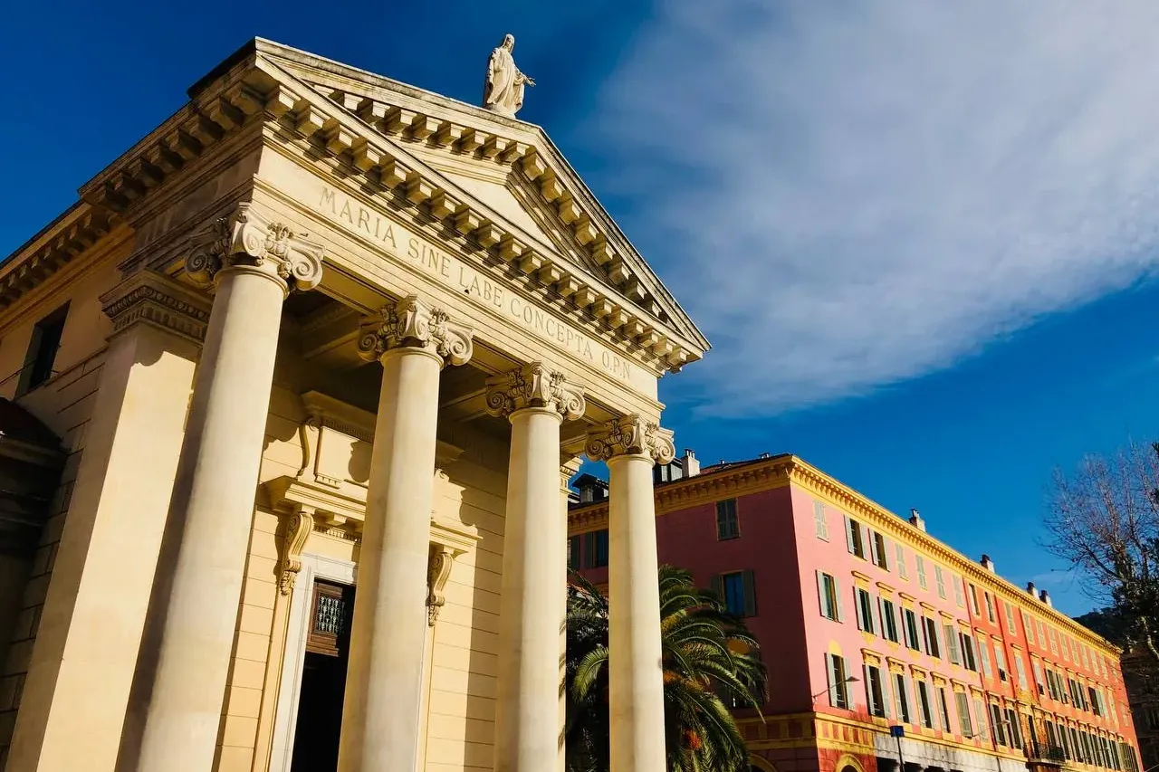Capilla neoclásica con columnas, frontón y estatua bajo cielo azul, junto a un edificio rosa con arcadas en Niza.