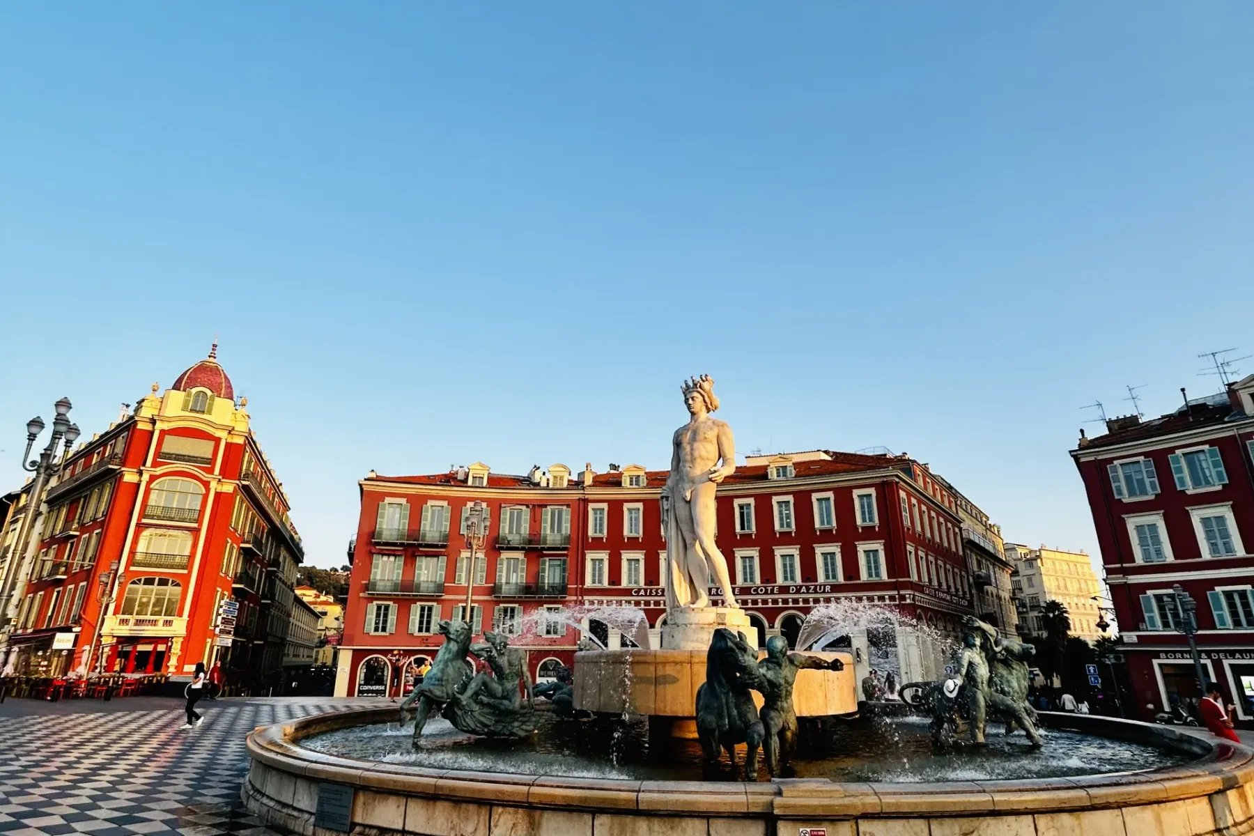 Fontanna Słońca (Fontaine du Soleil) w Nicei ze statuą Apolla i brązowymi postaciami, otoczona czerwonymi budynkami pod bł...