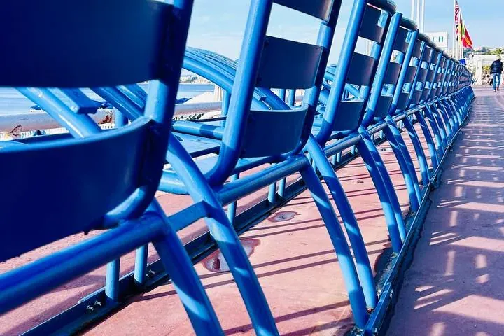 Una fila de icónicas sillas azules en el Paseo de los Ingleses de Niza, mirando al mar bajo un cielo azul claro. Las sombr...
