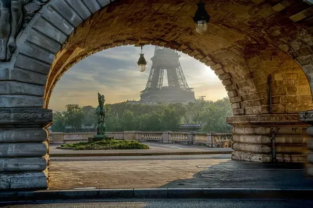 Eiffel Tower seen through a stone archway of a bridge, with a green statue and trees in the foreground under a bright sky.