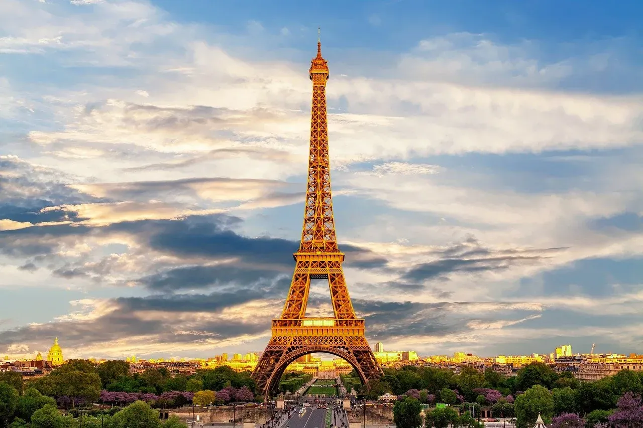 The Eiffel Tower rises above Paris at golden hour, with city buildings, green trees, and scattered clouds in the sky.