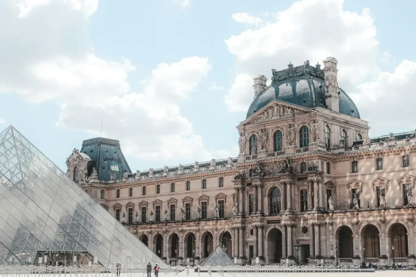 The Louvre Museum palace building with a dark dome and the glass pyramid in the foreground under a cloudy sky.