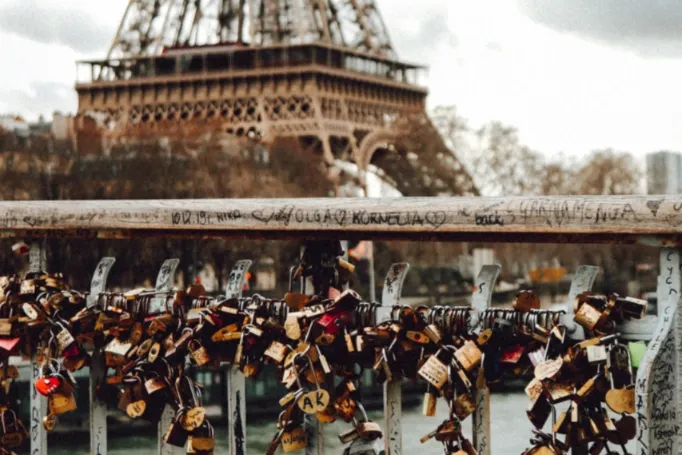 Numerous 'love locks' of various sizes and colors are attached to a grey metal bridge railing. The Eiffel Tower is blurred...