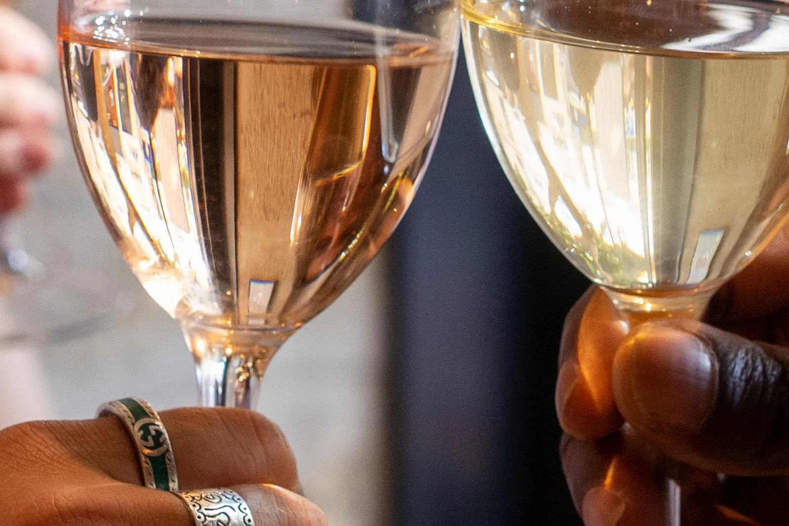 Zwei Hände halten Weingläser, eines mit Roséwein und eines mit Weißwein, die beim Anstoßen klingeln. Eine Hand trägt Ringe.