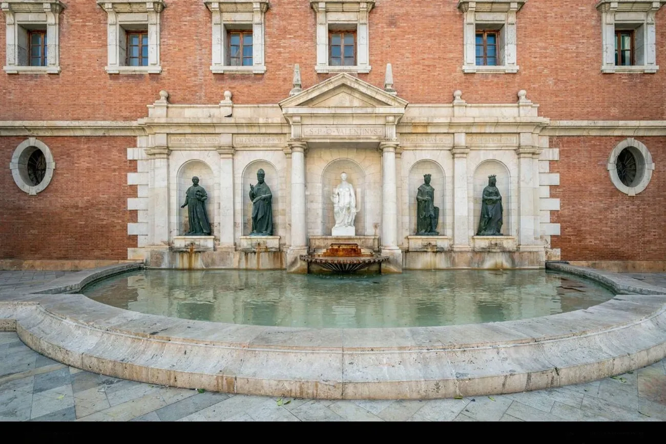 Una ancha fuente de piedra curvada frente a un edificio de ladrillo con estatuas en hornacinas.