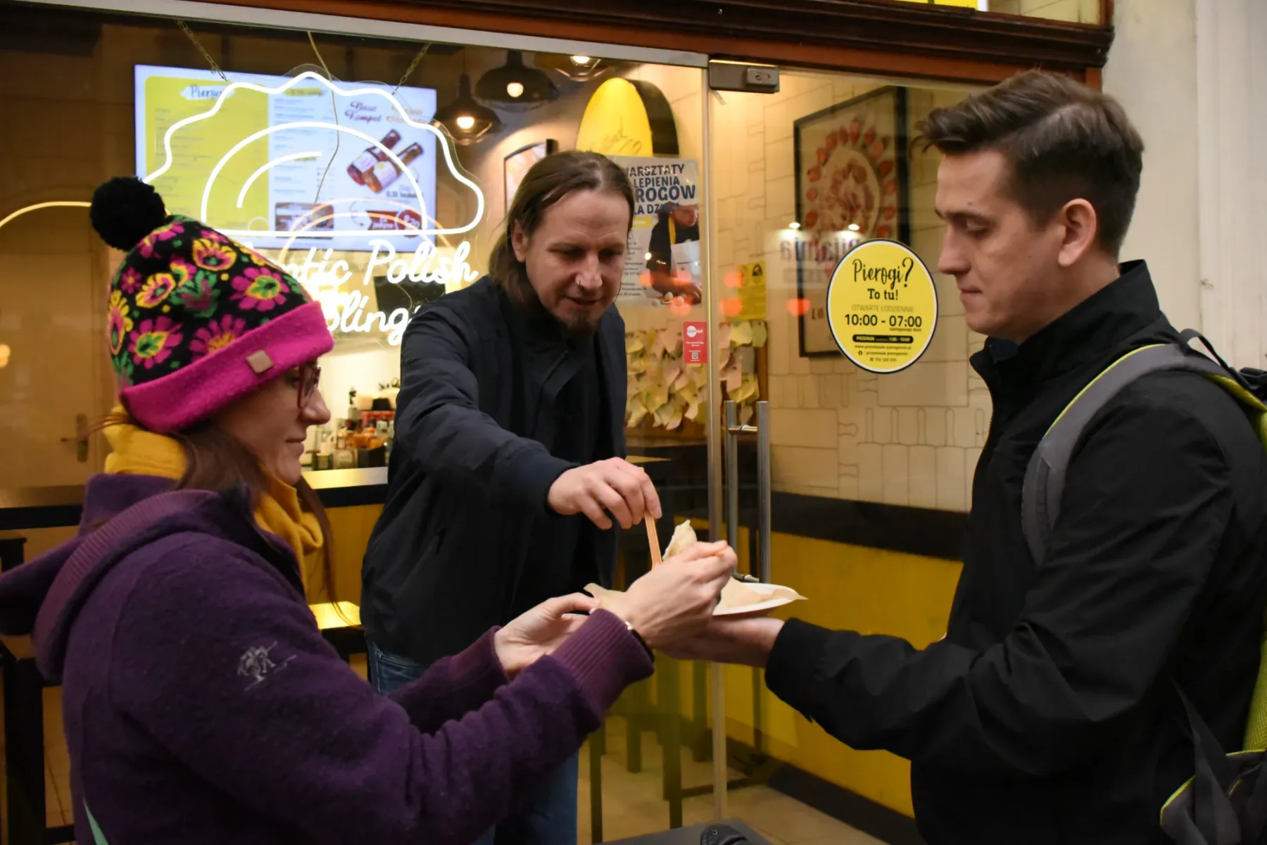 Three people standing outside a dumpling shop in Krakow, sharing pierogi from a paper plate.