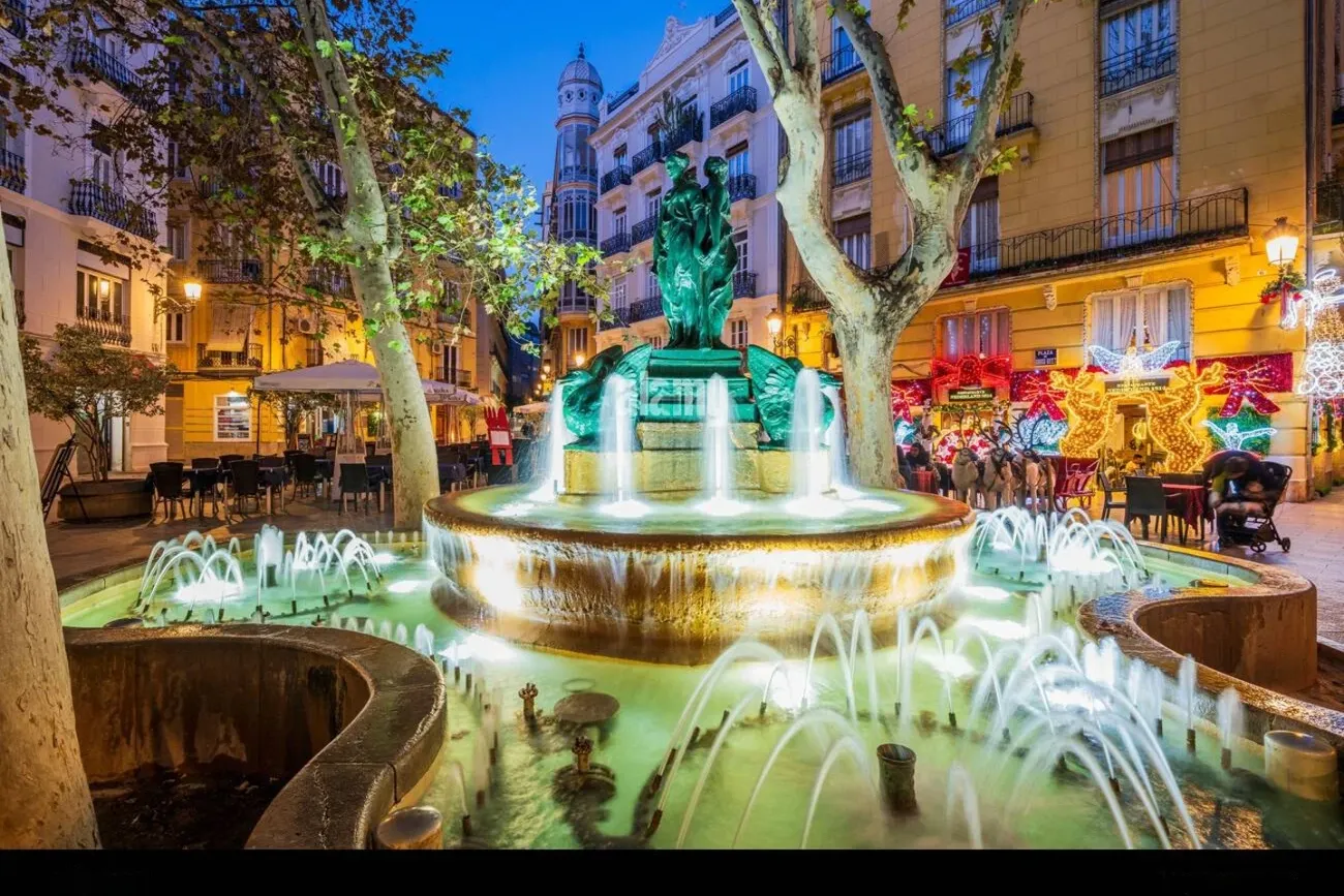 Una fuente iluminada en la Plaza Rodrigo Botet de Valencia, rodeada de árboles y edificios con luces de Navidad.