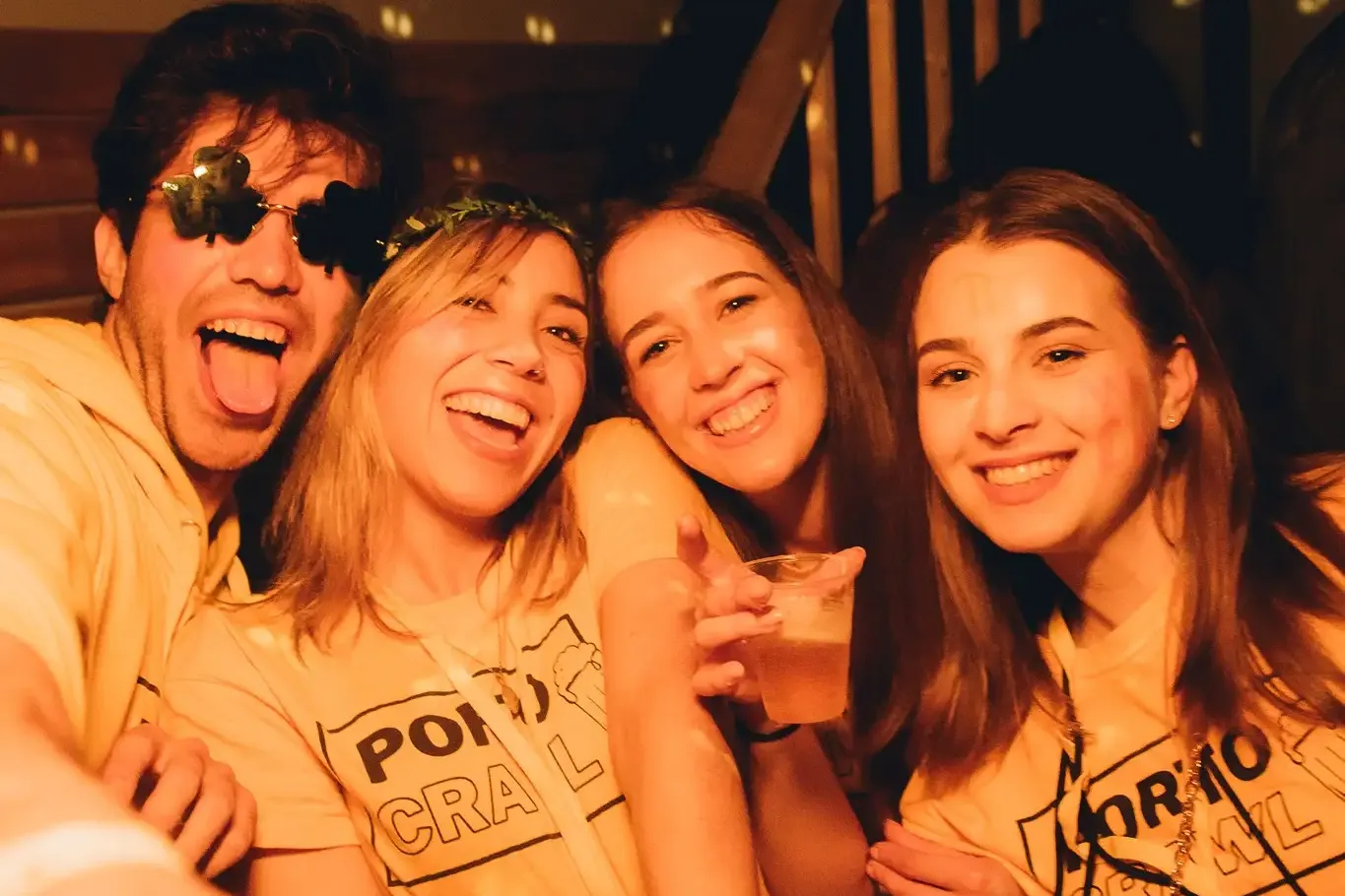 Vier junge Leute, zwei Männer und zwei Frauen, lächeln in einer Bar. Sie tragen gelbe 'PORTO PUB CRAWL' T-Shirts und halte...