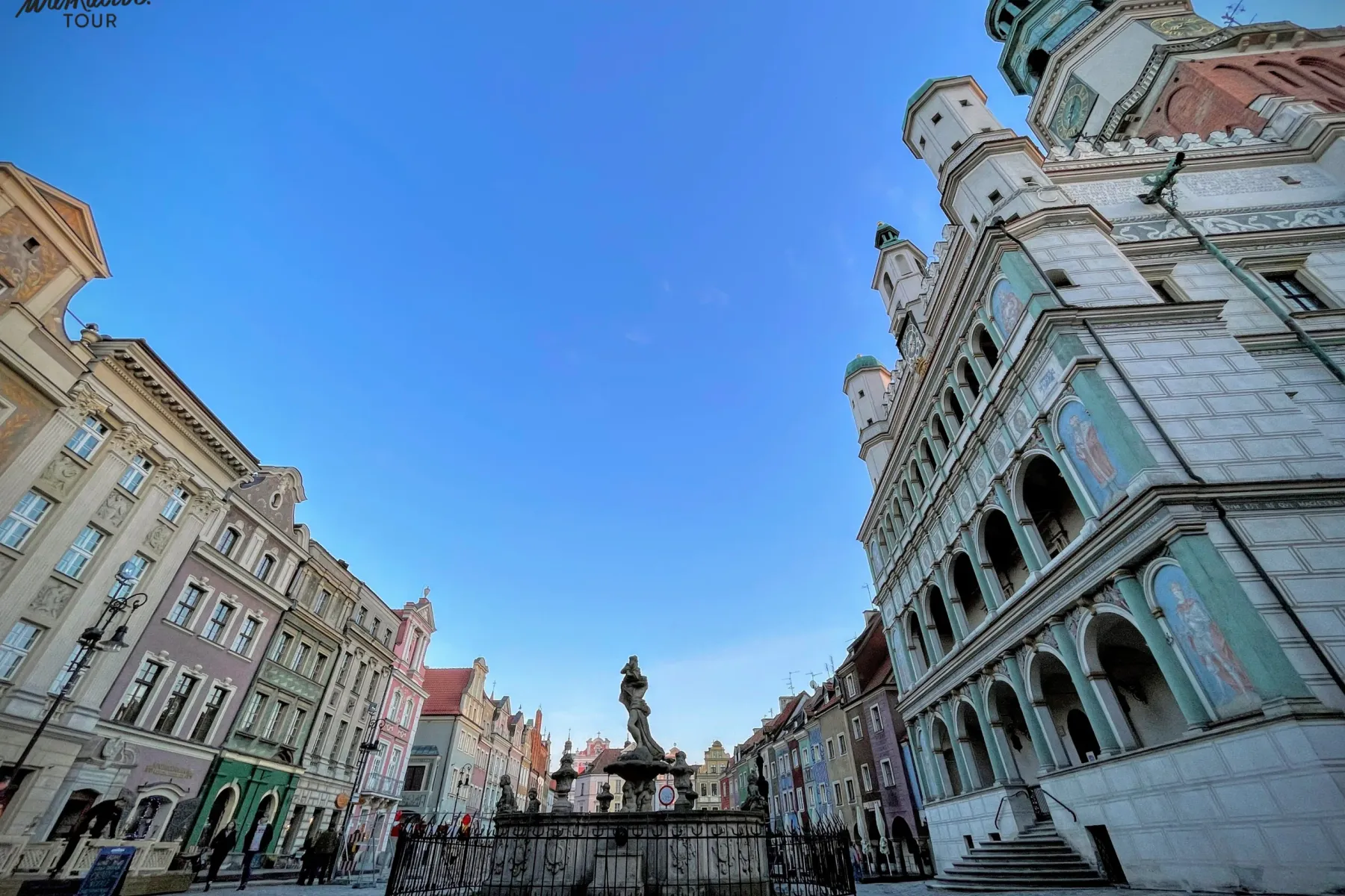 Panoramic view of Poznań Old Market Square with colorful tenement houses, a large fountain, and the ornate Renaissance Tow...
