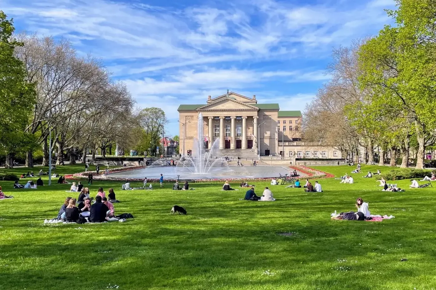 Grand Theatre building, fountain, and people relaxing on green grass in Poznań park on a sunny day.
