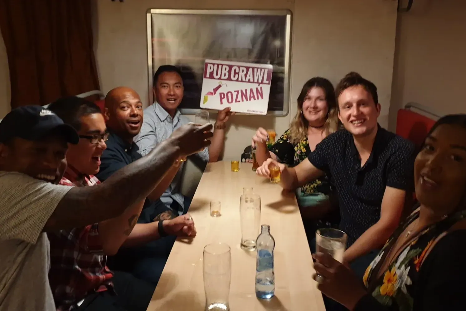 Diverse group of young adults smiling at a table in a pub, some holding drinks. One person displays a 'Pub Crawl Poznan' s...