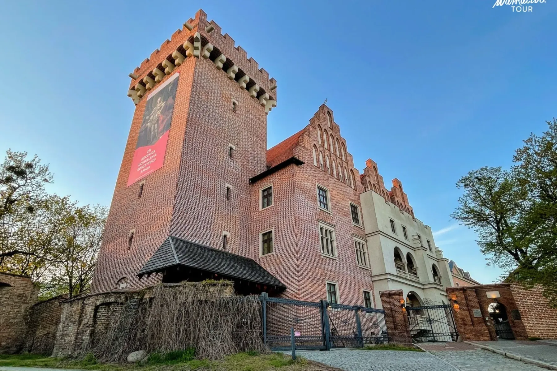 Tall brick castle with a prominent tower and medieval architecture, set against a clear blue sky. A cobbled path leads to ...