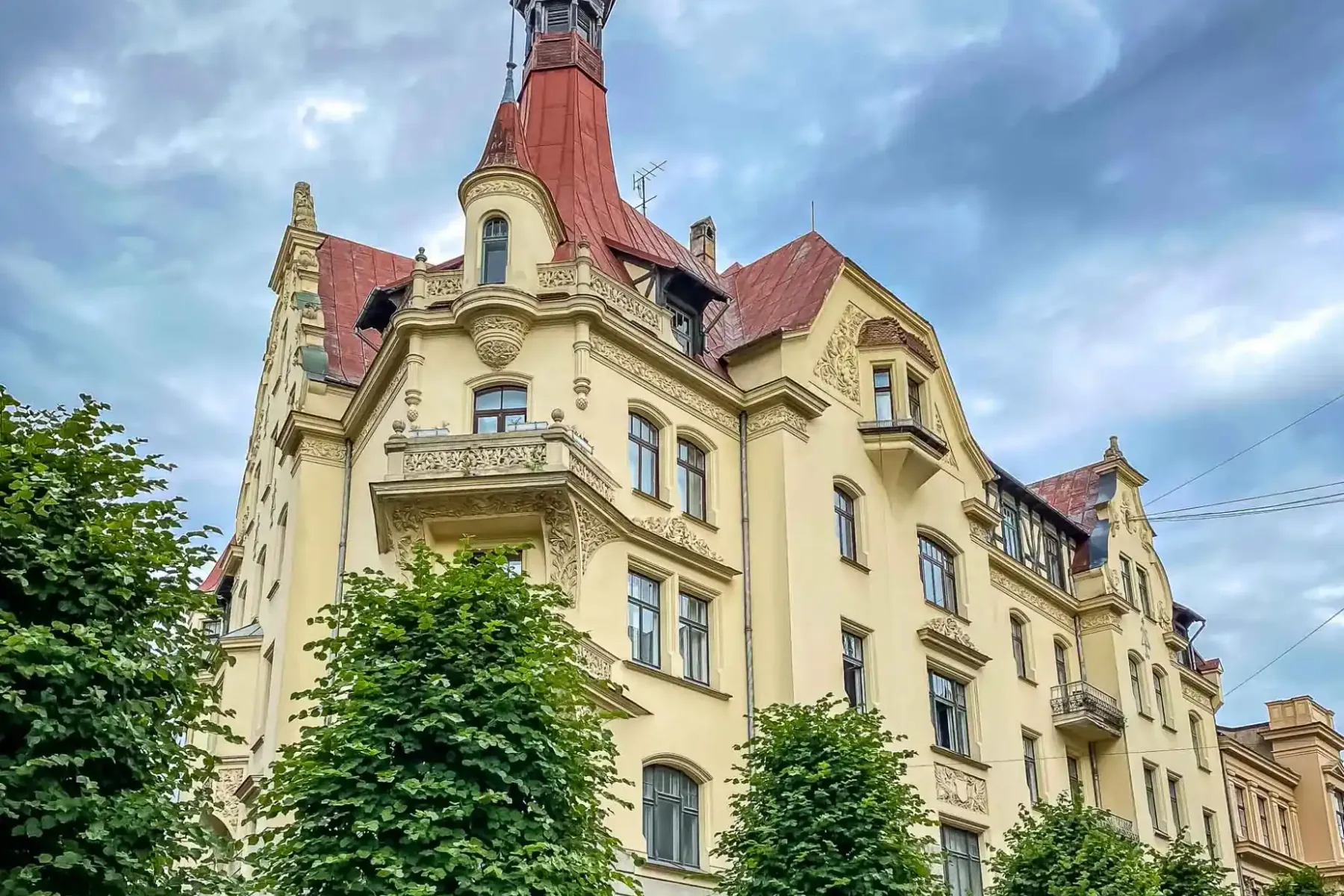 Verziertes Jugendstilgebäude in Riga mit gelber Fassade, rotem Dach und Turm, umgeben von grünen Bäumen vor bewölktem Himmel.