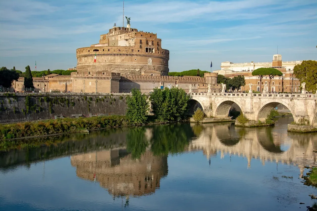 Die Engelsburg und die Engelsbrücke über dem Tiber, mit Spiegelungen im Wasser unter blauem Himmel.