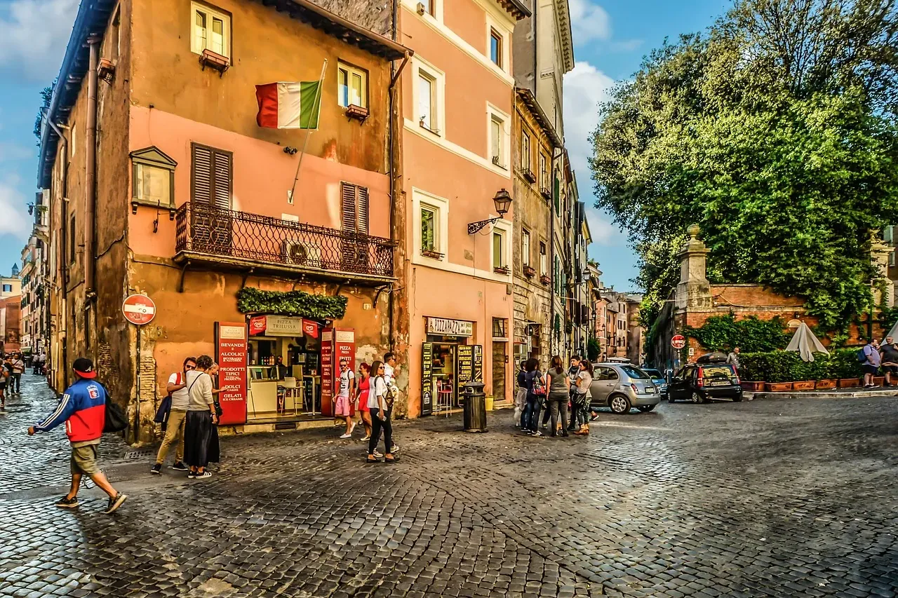 Calle adoquinada en Roma con edificios históricos, bandera italiana, personas caminando y coches aparcados bajo un cielo a...