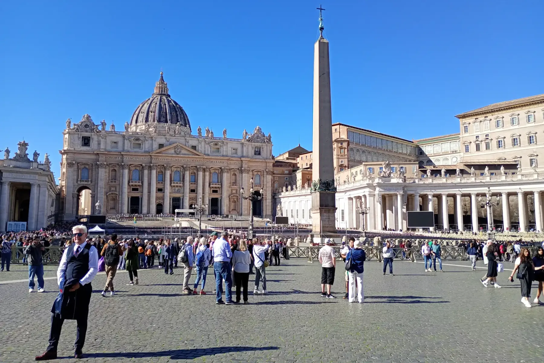 Tourists walking in St. Peter's Square with the central obelisk and St. Peter's Basilica facade behind.