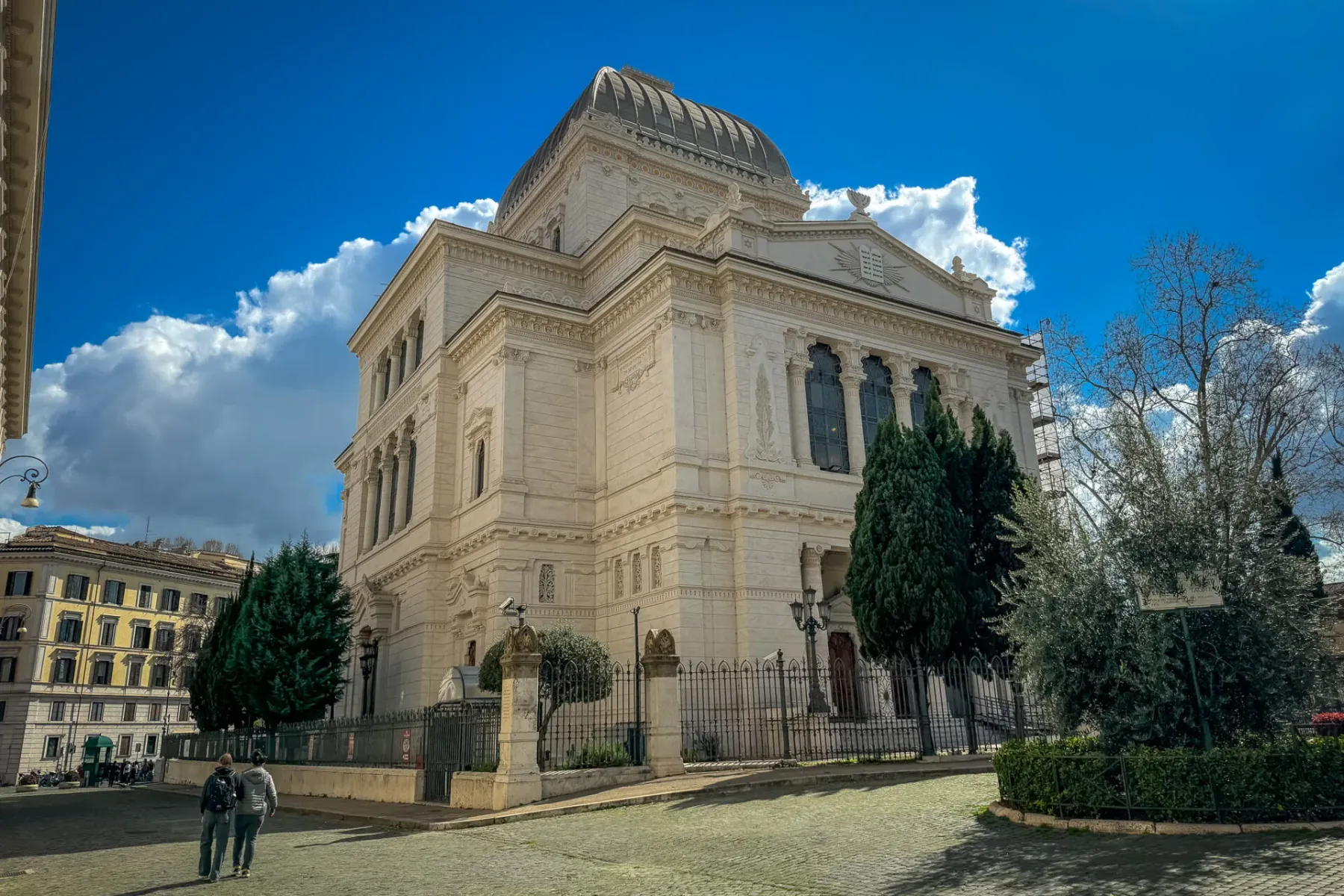 Die Große Synagoge von Rom mit Kuppel, eine Kopfsteinpflasterstraße, grüne Bäume und zwei Personen unter blauem Himmel.