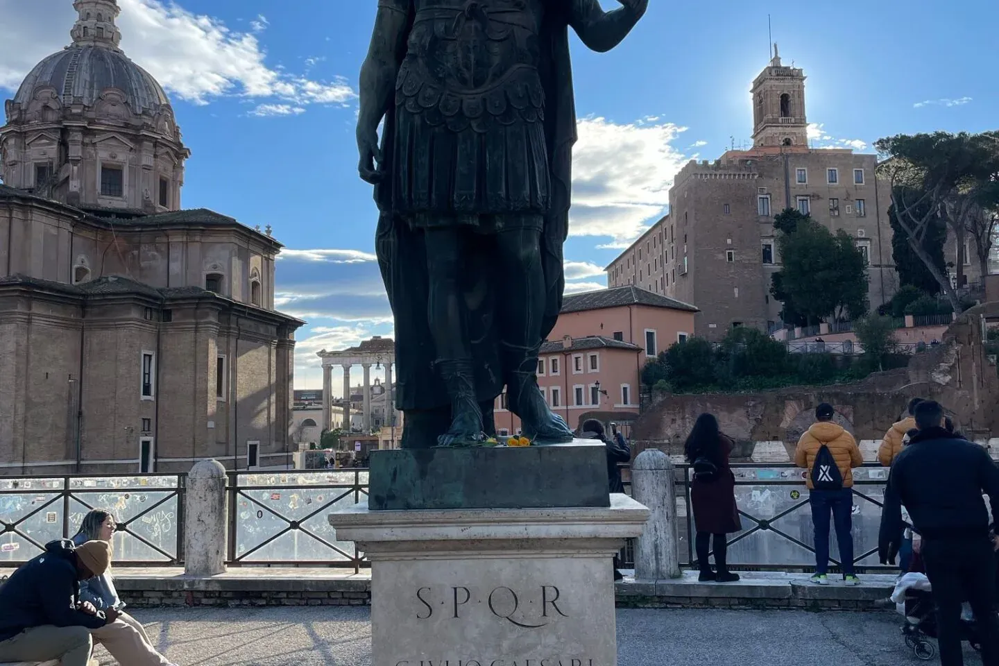 Bronzestatue von Julius Cäsar in Rom, mit Ruinen des Forum Romanum und einer Kuppelkirche unter blauem Himmel.
