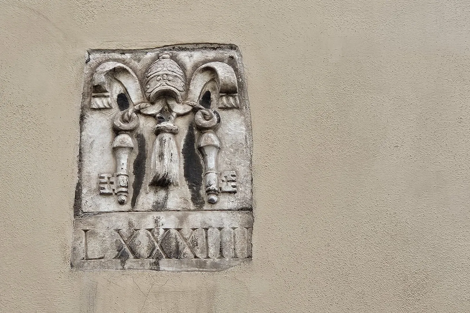 Steinrelief an einer strukturierten Wand, das eine Papsttiara, gekreuzte Schlüssel, Quasten und römische Ziffern LXXXIII z...